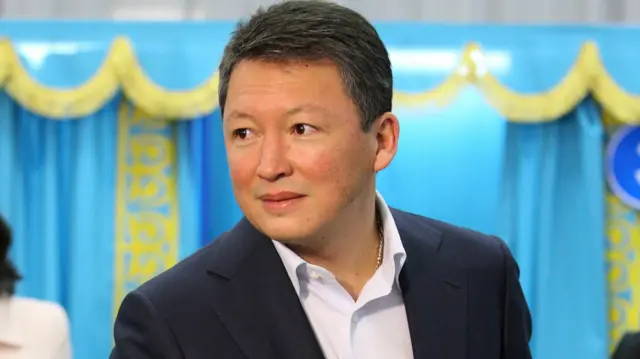 Тимур Кулибаев (на фото 2011 года) в костюме и рубашке без галстука на фоне избирательных кабинок цветов казахстанского флага