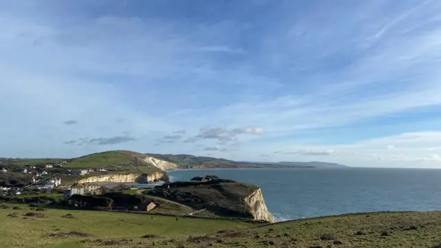 Vista de la costa con un cielo soleado y brillante, con solo algunas nubes de buen tiempo. Se pueden ver zonas de hierba sobre un mar azul.