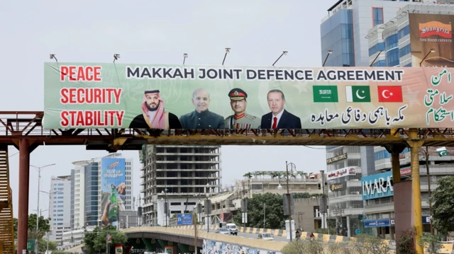 Una valla publicitaria sobre el tráfico muestra a los líderes de Turquía, Arabia Saudita y Pakistán tras la firma del Acuerdo Conjunto de Defensa de La Meca, en Karachi. Debajo se ven coches y personas.