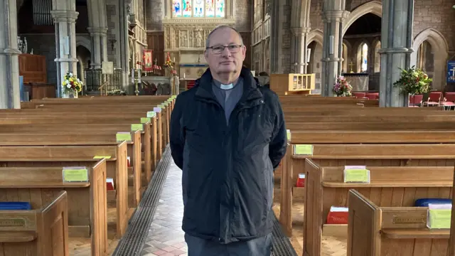 El reverendo Martin Jacques, vicario de la iglesia de San Pedro en Budleigh Salterton, aparece de pie en el pasillo de la iglesia con las manos a la espalda. Lleva un abrigo negro sobre una camisa gris y un cuello de vicario blanco debajo. Tiene el pelo corto y canoso y lleva gafas.