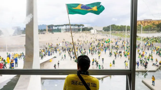 Uma pessoa vestida com uma camiseta amarela está de costas, onde se lê "Deus acima de tudo", olhando pela janela. Lá fora, manifestantes participam do ato antidemocrático, que pedia por um golpe de Estado, em 8 de janeiro de 2023.