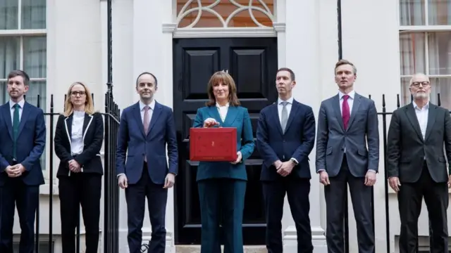 スーツを着た男女が7人、屋外に横並びで立っている。中央の青いパンツスーツの女性が、赤い箱型のかばんを胸元に掲げている。背後には白い建物と黒い鉄柵、黒い玄関のドアが写っている