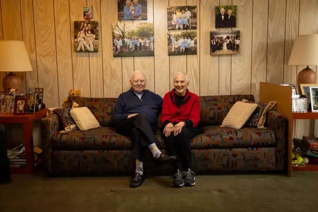 Ralph Rehbock sentado en un sofa oscuro, con cojines, junto a su esposa en la sala de su casa. Atrás, en la pared, se pueden ver fotos familiares así como en una mesa que está al lado