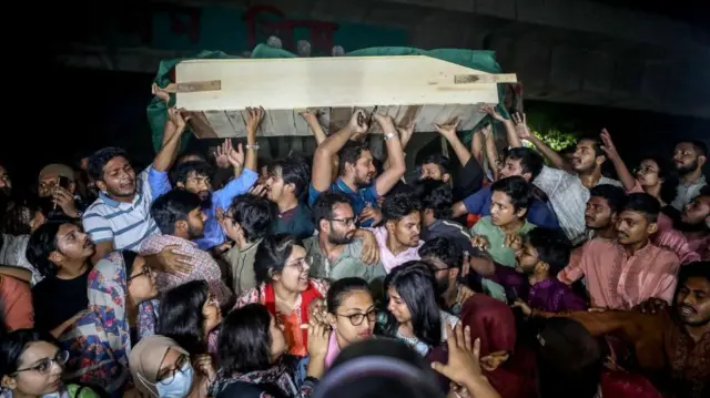 A tight crowd of people hold up a wooden coffin