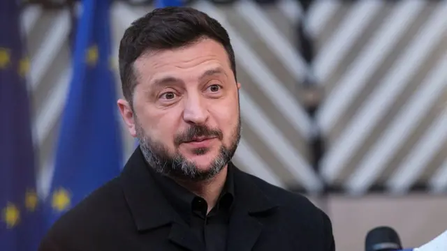 Prezydent Ukrainy Wołodymyr Zelenski - mężczyzna w średnim wieku, z ciemnymi włosami, piwnymi oczami i siwiejącą brodą - stoi przed mikrofonem. Ma uniesione brwi i wyraźnie zdumiony lub poruszony wyraz twarzy. 