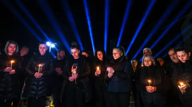 Седам жена у црном са свећама у рукама, плави сноп светлости се види у даљини 