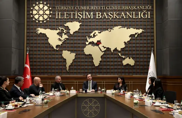 Fahrettin Altun, bir masanın başında oturuyor. Arkasında İletişim Başkanlığı yazıyor. 