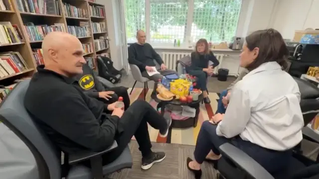 白いジャケットを着た女性が、黒い衣服の男女と車座になって話し込んでいる