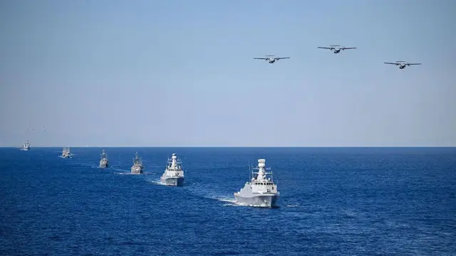 6 adet savaş gemisi denizde ufka doğru küçülerek sıralanmış, onları takip eden üç savaş uçağı da havada sıralanmış.