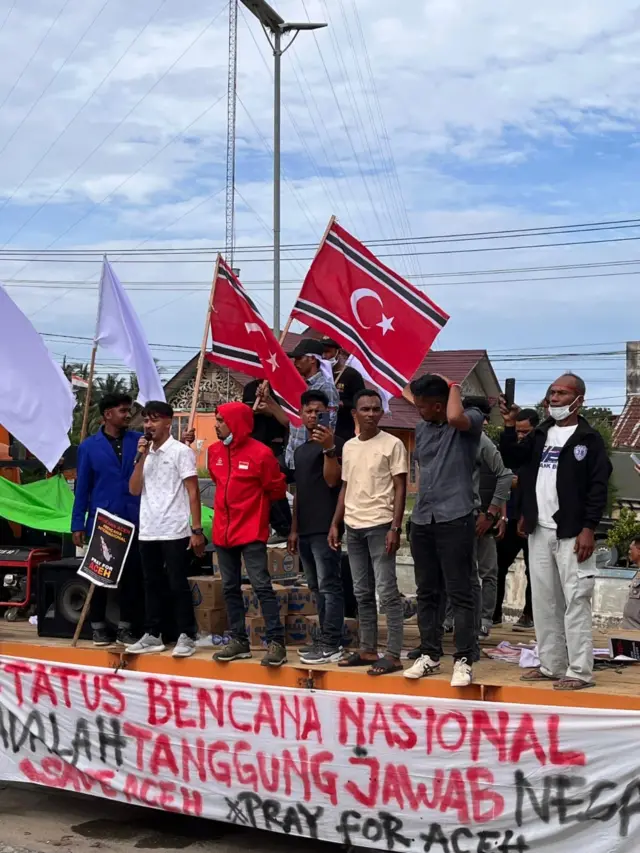 Peserta aksi damai di depan Kantor Bupati Aceh Utara pada Kamis (25/12), mengibarkan bendera putih dan bendera bulan bintang di atas sebuah panggung kecil sambil berorasi. Di bagian depan panggung terdapat tulisan "Status bencana nasional adalah tanggung jawab negara #saveaceh #prayforaceh.