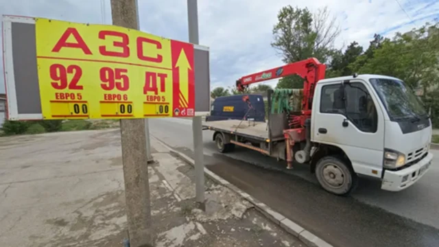 Табло з цінами на пальне біля автозаправної станції. На тлі — вантажівка з краном на міській вулиці.