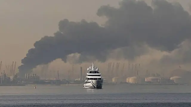 تصویر یک کشتی در بندر جبیل علی دبی. دود ناشی از انفجار در شهر دبی از دور نمایان است. 