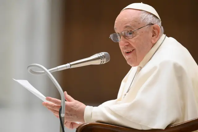 Papa Francisco sorri, sentado em frente ao microfone