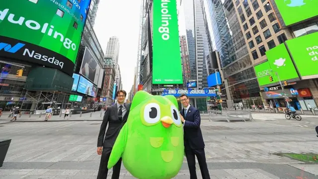 A foto foi feita na rua, com prédios ao redorcassinos famososdois homens jovens vestidos com terno e gravata e uma coruja verde, símbolo do Duolingo, no meio deles. 