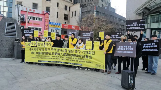 一群身穿黑色与黄色服装的人手持标语牌和一幅大型横幅；横幅以韩文和英文写着：“呼吁中国政府停止将朝鲜人遣返回朝鲜的记者会”。

