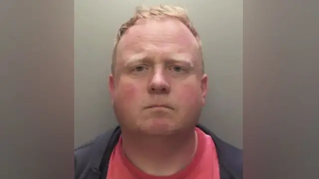 Robert Bush wit short, light brown hair show for dis straight‑on mugshot foto. Im no dey smile or frown, im wear dark jacket ontop red shirt. Di background dey plain and grey.