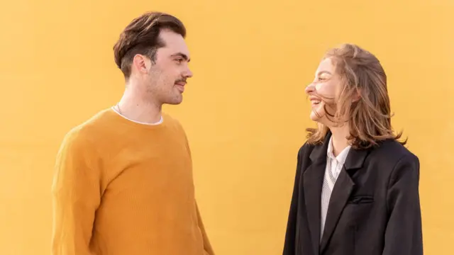 Młody mężczyzna i kobieta śmieją się i patrzą na siebie, stojąc na tle intensywnie żółtej ściany.