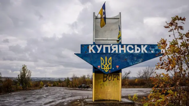 Знак на въезде в город Купянск. Знак синего цвета с большими белыми буквами «Купянск» и желтым трезубцем посередине. Над знаком прикреплен украинский флаг, поврежденный и немного разорванный.