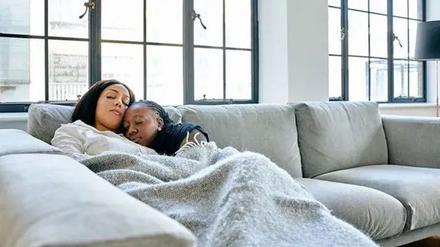 Дві дівчини лежать на дивані у віталі накрившись пледом