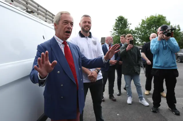Nigel Farage, lider partii Reform UK, przemawia z uniesionymi rękami, stojąc obok białego samochodu dostawczego. Za nim stoi lekko uśmiechnięty kandydat partii na deputowanego z okręgu Makerfield, Robert Kenyon. Za nimi, po prawej stronie zdjęcia, stoi grupa mężczyzn. Jeden z nich trzyma w górze kamerę wideo z dołączonym mikrofonem. Inny trzyma w górze smartfon. 