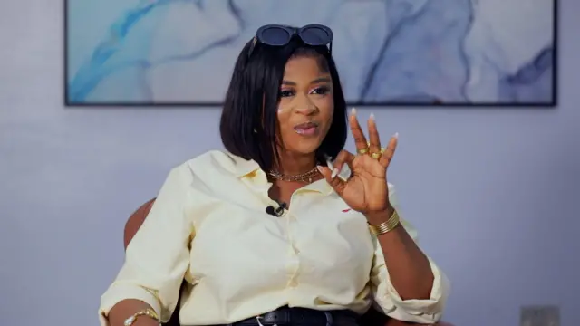 Imisioluwa Ayanwale to se ipo kinni nile Big Brother Naija fun ọdun 2025 joko lori aga kan, o na ọwọ osi soke bii ẹni to n ta ika, o fi irun wiigi dudu sori, o si fi igo akọ soke sori irun rẹ, o wọ asọ shaat alawọ miliki, o ọ sokoto alawọ buluu, to si lo bẹliti sori sokoto naa.