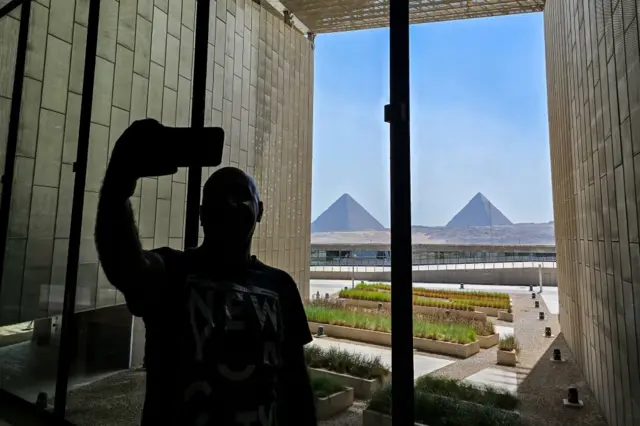 Pessoa tira selfie dentro do museu, com pirâmides ao fundo vistas através4kasinovidro 