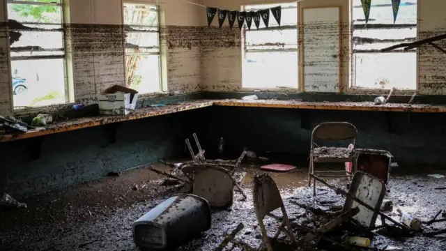 「WELCOME（ようこそ）」とペナントがつるされた部屋。壁の上部に泥が平らな線となって残り、浸水がどこまで達したか推察できる。その下のカウンターテーブルや床、床に散らばるパイプ椅子などはいずれも泥まみれの状態