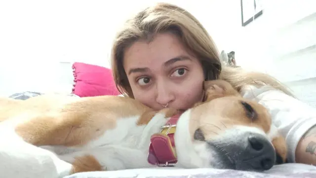 Catalina recuesta su cabeza sobre un perro que está dormido sobre una cama y mira a la cámara