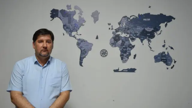 مردی با پیراهن آستین کوتاه کنار نقشه جهان روی دیوار ایستاده به دوربین نگاه می‌کند 
