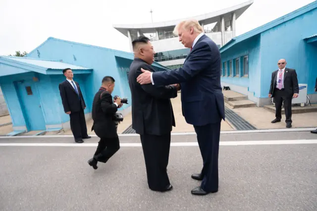 Trump e Kim Jong-un se cumprimentando em pátio, com seguranças atrás