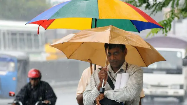වර්ෂාව හේතුවෙන් කහ පැහැති කුඩයක් ඉහලාගෙන ගමන් කරන පුද්ගලයෙක්, වැස්සේ තෙමෙමින් යතුරු පැදියක් ගමන් කරන අයෙක් පසුබිමින් දිස්වේ 