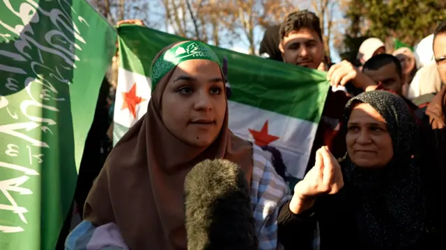 Leyla yanında akrabasıyla kameraya konuşuyor, arkasında Suriyeli muhaliflerin bayrakları var.