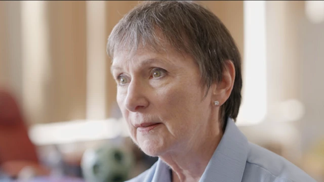 Lee McKenzie is a woman in her 60s with short greying dark hair. She is wearing a light blue shirt with a collar. Her head and shoulders can be seen. She is looking off to the left of the picture.