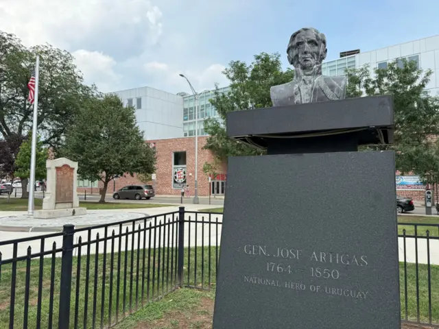 Busto del general José Artigas, prócer de Uruguay, en la plaza frente al ayuntamiento de Elizabeth. A la izquierda se ve un mástil con una bandera de Estados Unidos y, de fondo, la fachada de una escuela secundaria.