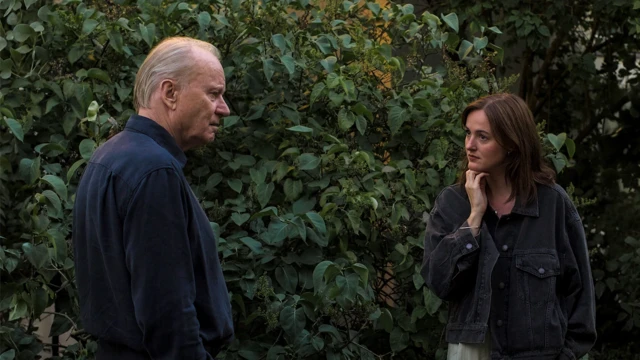 Чоловік і жінка стоять просто неба перед щільними зеленими кущами. Обидвоє вдягнені у темний одяг і стоять одне навпроти одного, ніби спілкуються.