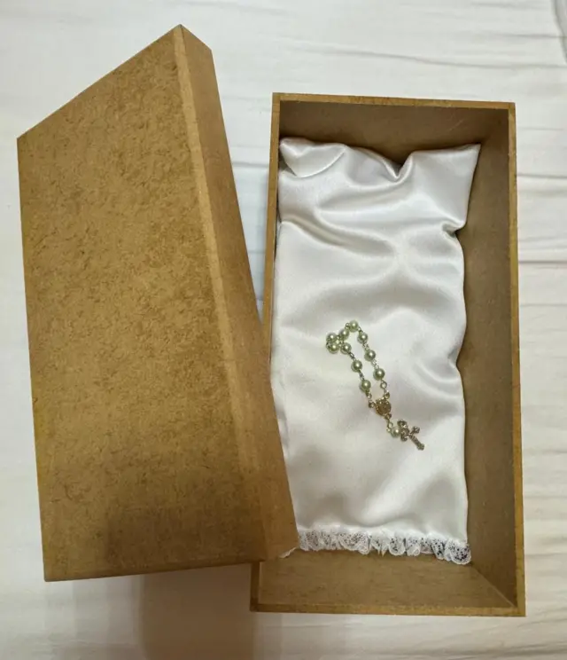 Foto de uma caixa de madeira menor que uma caixa de sapatos, aberta, contendo um pequeno terço sobre uma almofada branca  