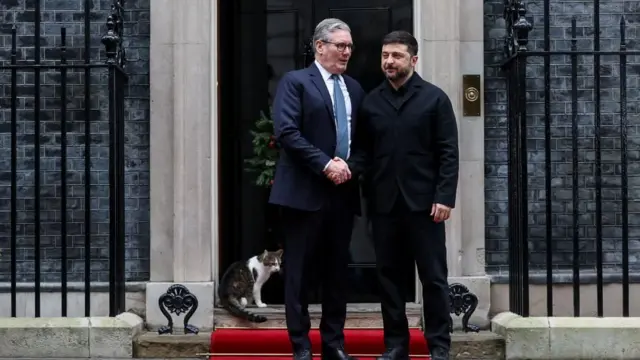 Brytyjski premier Keir Starmer ściska dłoń prezydenta Ukrainy, Wołodymyra Zełenskiego, przed wejściem do domu przy Downing Street 10. Za nimi, na progu siedzi Larry. 