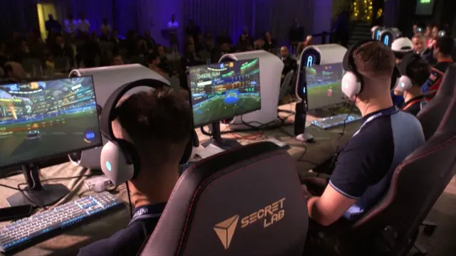 Kilku mężczyzn siedzi na krzesłach, grając w gry komputerowe na dużych ekranach. Mają na uszach słuchawki i trzymają kontrolery. W tle widać publiczność.