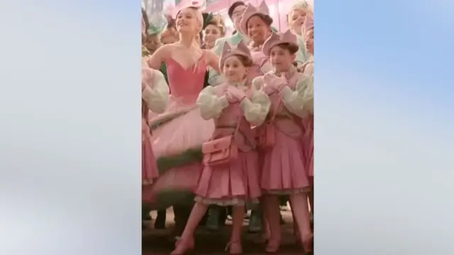 Captura de pantalla de Ariana Grande rodeada de un grupo de chicas vestidas con trajes rosas. Lily aparece en primer plano.