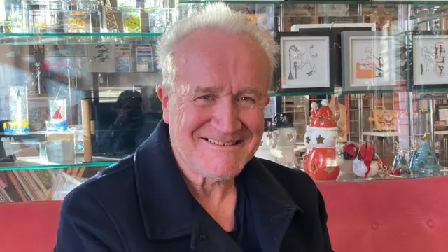 Mike Padgham, propietario de la residencia de ancianos Santa Cecilia, es un hombre mayor, de pelo corto y blanco, barba incipiente y abrigo negro. Está sentado en un sofá rojo.