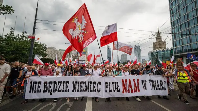 Уличная хода в центре Варшавы. Впереди колонны участники держат длинный белый баннер с черной и красной надписью на польском языке: «Не будет немец плевать нам в лицо». Над толпой развеваются многочисленные польские флаги, а также большие красные знамена с белым орлом.