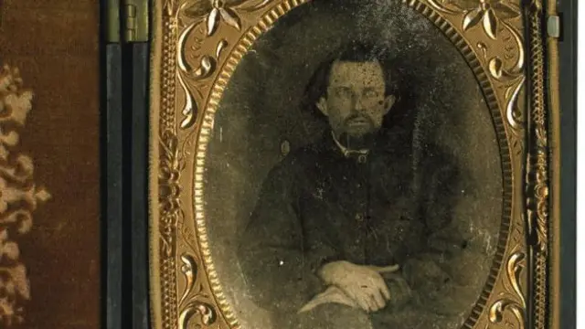 A imagem mostra um antigo retrato fotográfico em tom sépia, emoldurado por uma moldura dourada ornamentada. No centro, aparece um homem de expressão séria, com barba e cabelos escuros, vestido com roupas escuras formais da época. É Nathan Bedford Forrest.