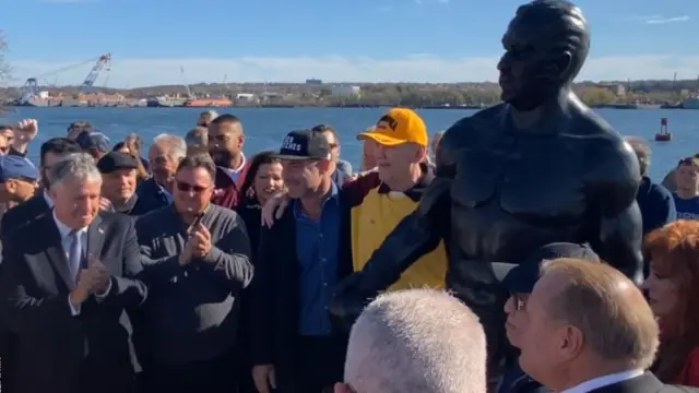 Chuck Wepner posa com gorro amarelo junto à estátua construída em sua homenagem, no Estado americano de Nova Jersey.