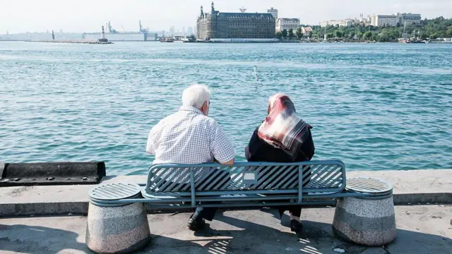 Kadıköy'de Haydarpaşa Garı'nı gören bir açıdan deniz kenarındaki banka oturmuş yaşlı bir çift. Arkadan gördüğümüz çiftten soldaki erkek olanı üzerinde kısa kollu bir gömlekle otururken sağdaki kadının üstünde ise siyah bir kıyafet ve desenli bir başörtüsü var