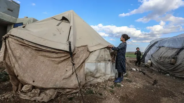 Une fille se tient à côté d'une tente fabriquée sommairement - l'un des côtés est maintenu en place par de petits rochers, province d'Idlib, mars 2024.