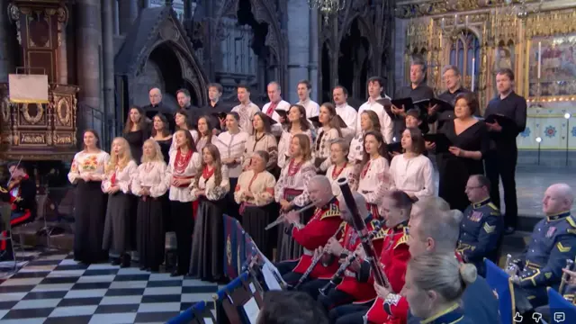 Інтер’єр великого готичного собору з декоративним золотистим вівтарем на задньому плані. У центрі стоїть хор у кілька рядів, багато учасників одягнені у традиційні українські вишиванки. На передньому плані сидить оркестр у червоній формі з синіми та золотими деталями, музиканти тримають духові інструменти.