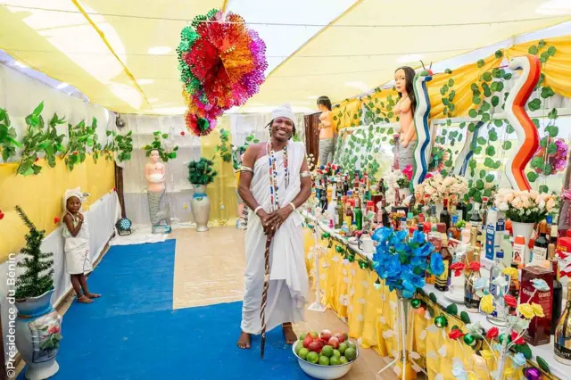 Un homme et un enfant en tissus blanc avec des perles autour du cou, debout dans un couvent avec des divinités.