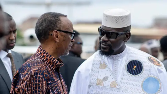 Vue de profil de Paul Kagame, vêtu d'une chemise africaine à motifs, discutant avec Mamady Doumbouya, vêtu d'un boubou blanc, d'un chapeau blanc et de lunettes noires.