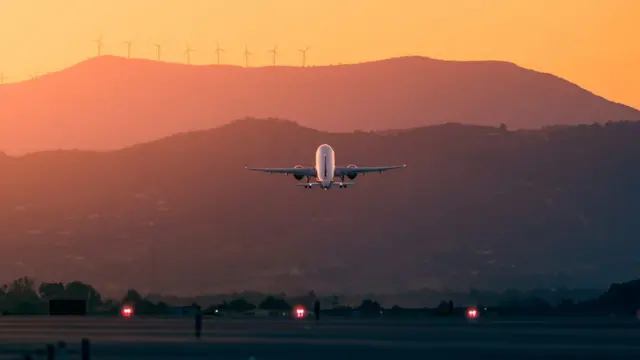 Avião decolando em dia de sol, com montanhas e turbinas eólicas ao fundo