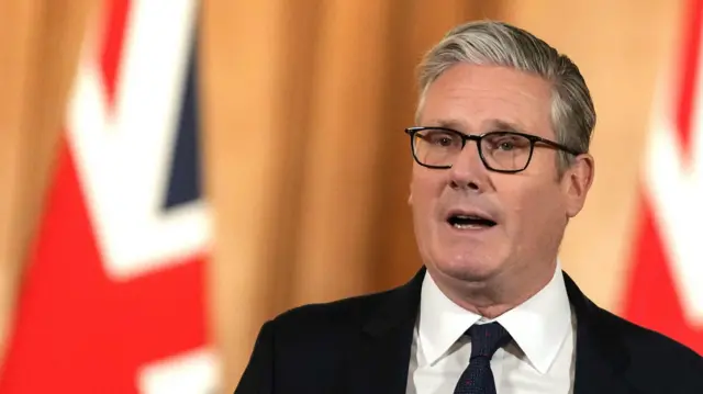 Keir Starmer veste terno, gravata e óculos durante um pronunciamento oficial, com expressão séria e a boca aberta no momento do discurso. Ao fundo, há bandeiras desfocadas do Reino Unido.